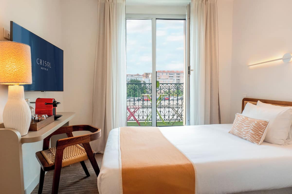 A hotel room with a bed, a desk with a lamp, a chair, and a view of the outside.