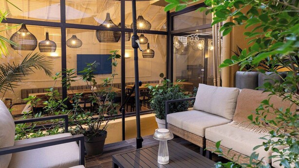 Un área de estar interior con pared de vidrio, varias lámparas colgantes y plantas en macetas.