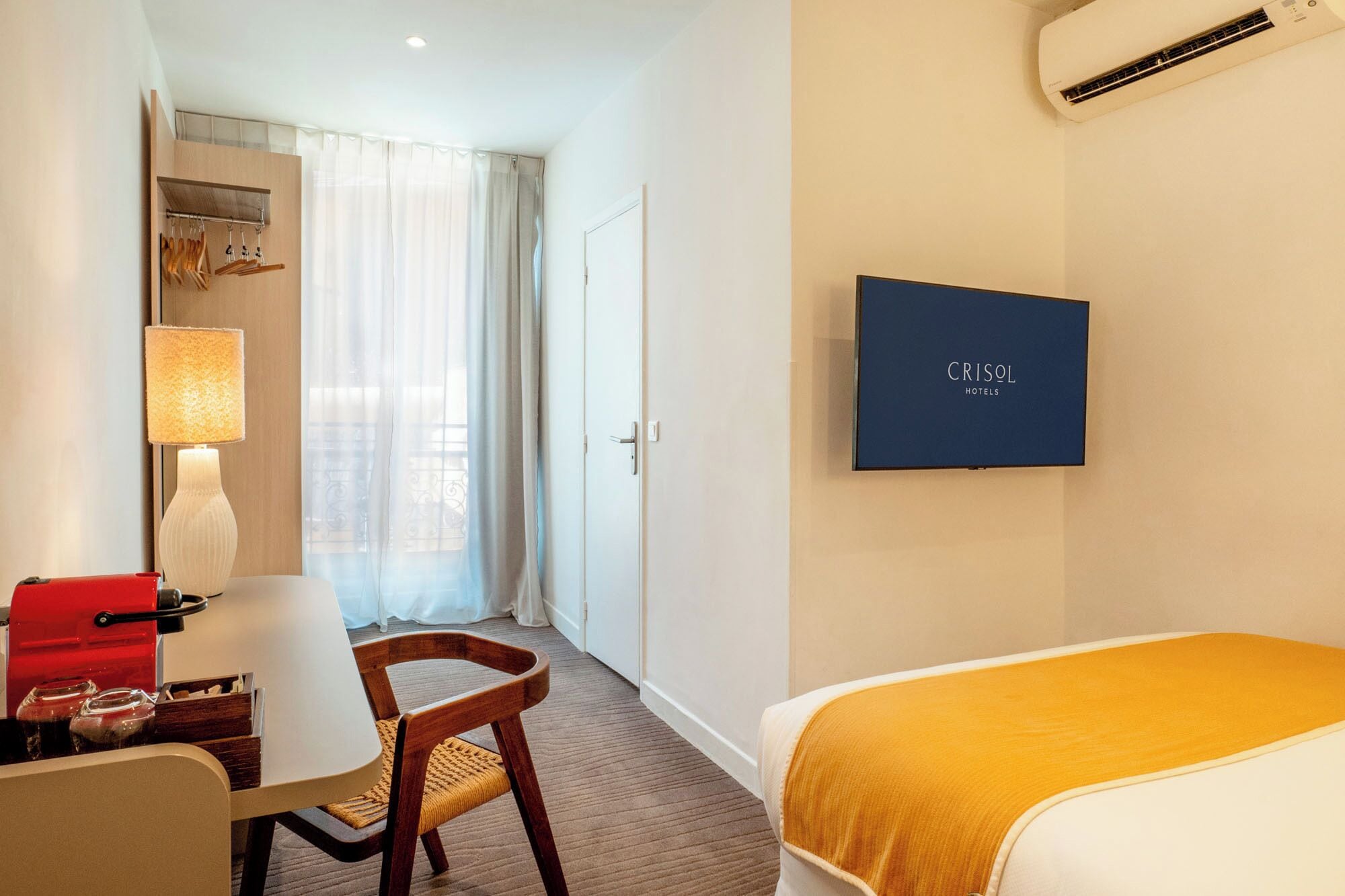 Habitación de hotel con cama, escritorio con lámpara, silla, televisor y ventana con cortinas.