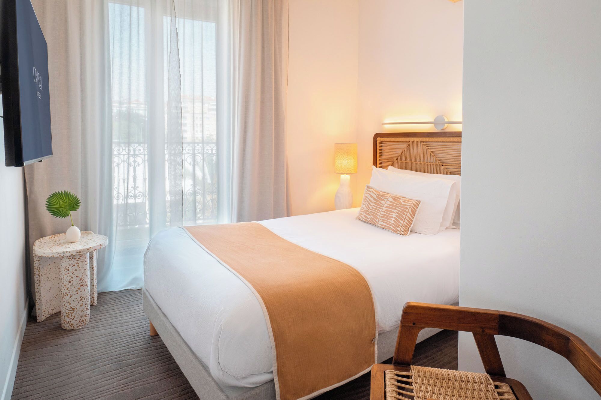 Quarto de hotel com cama, luminária de cabeceira, mesinha com uma planta e uma cadeira.
