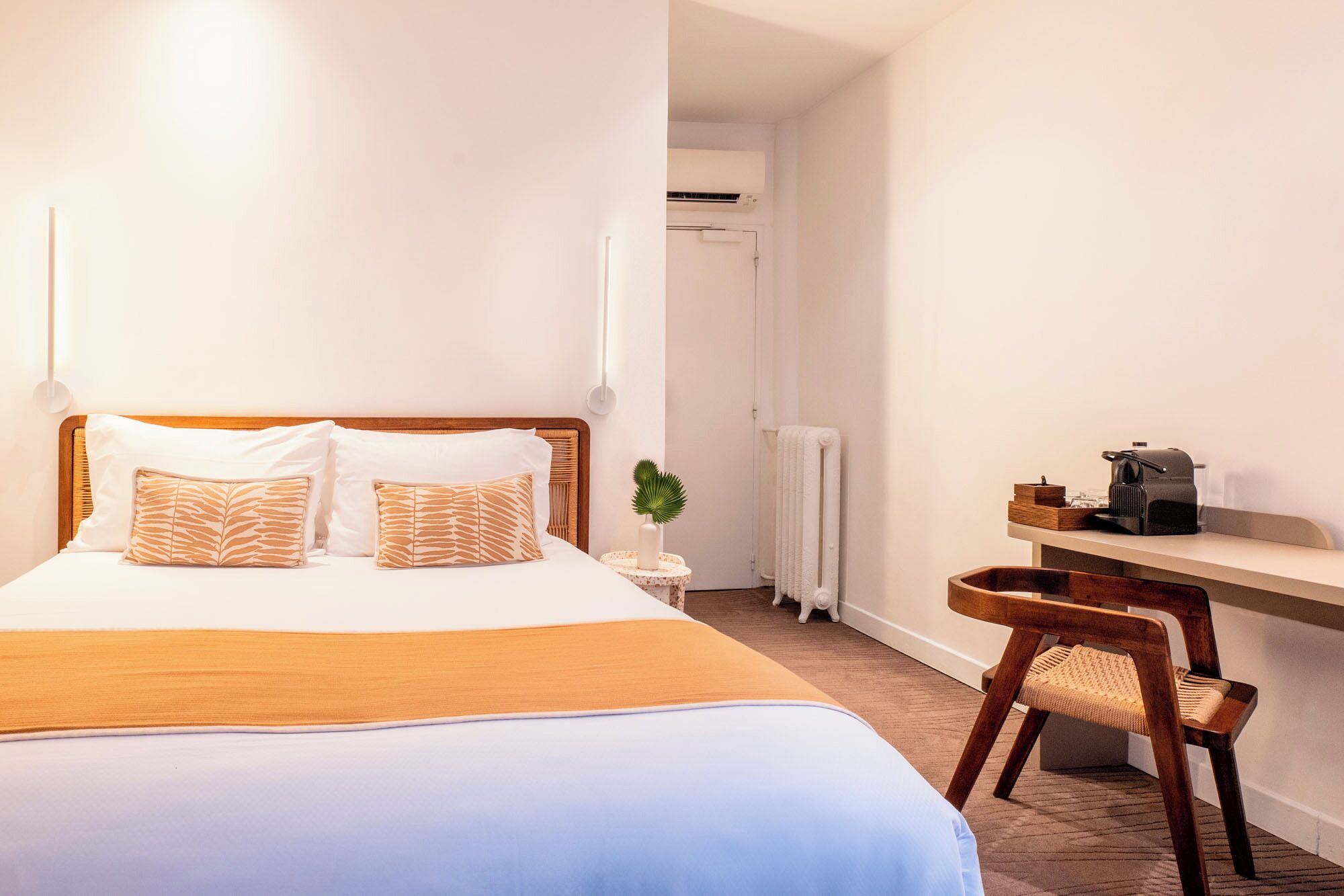 A hotel room with a bed, a desk, a chair, a radiator, and a small plant.