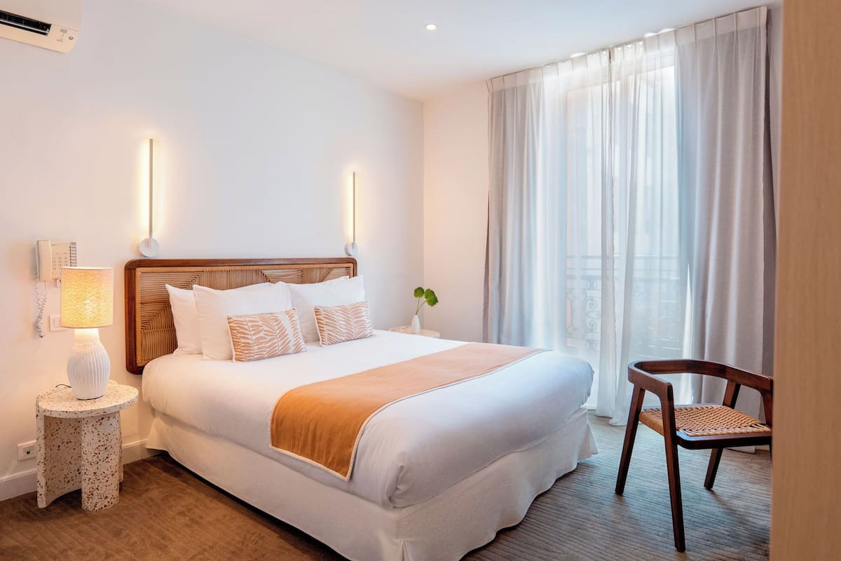 A hotel room with a large bed, bedside lamp, chair, and a window with sheer curtains.