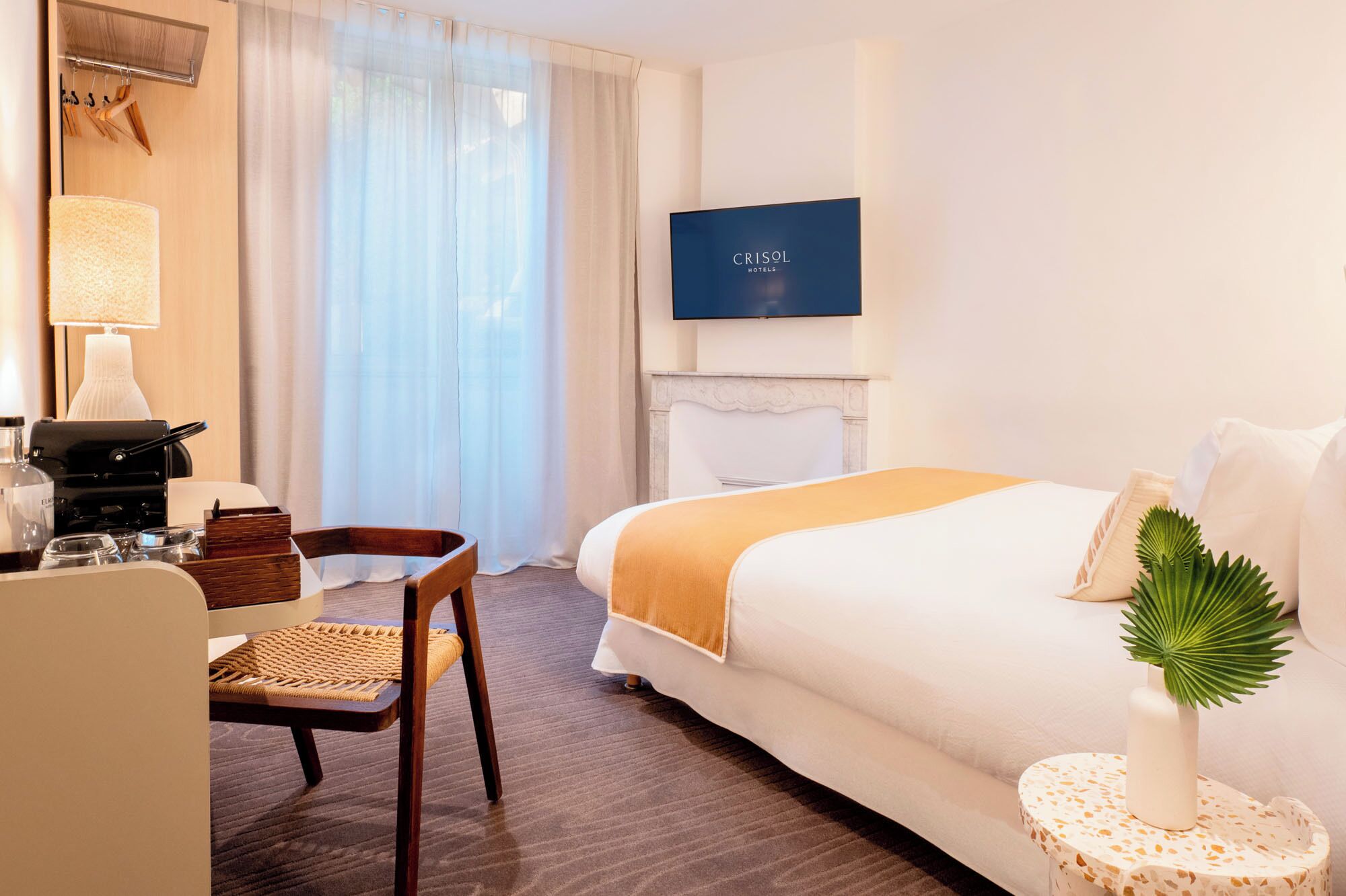 A hotel room with a bed, a TV, a desk with a lamp, a chair, and a small table with a plant.