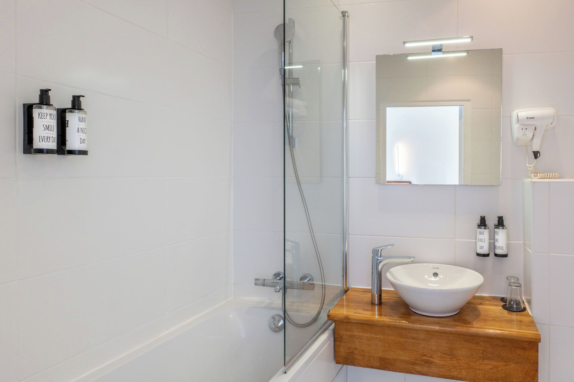 A modern bathroom with a glass-enclosed shower, a wooden vanity with a white sink, and two bottles of hand soap on the wall.