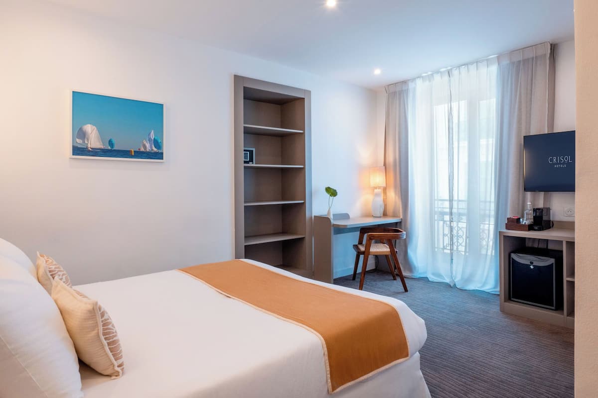 A hotel room with a bed, a desk, a chair, a TV, and a painting on the wall.