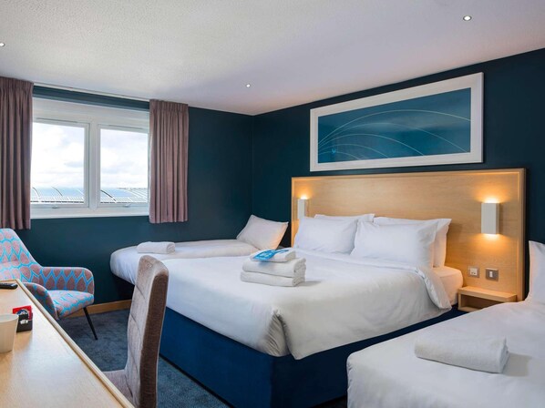 Ein Hotelzimmer mit einem großen Bett, einem Schreibtisch und einem Sessel. Es gibt ein Fenster mit Vorhängen, ein Bild an der Wand und eine Deckenleuchte.