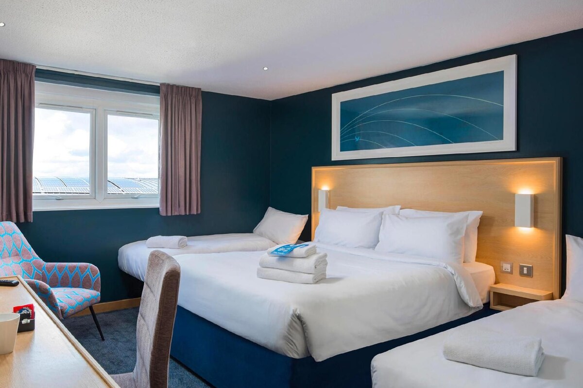 A hotel room with a large bed, a desk with a chair, a window with curtains, and a painting on the wall.