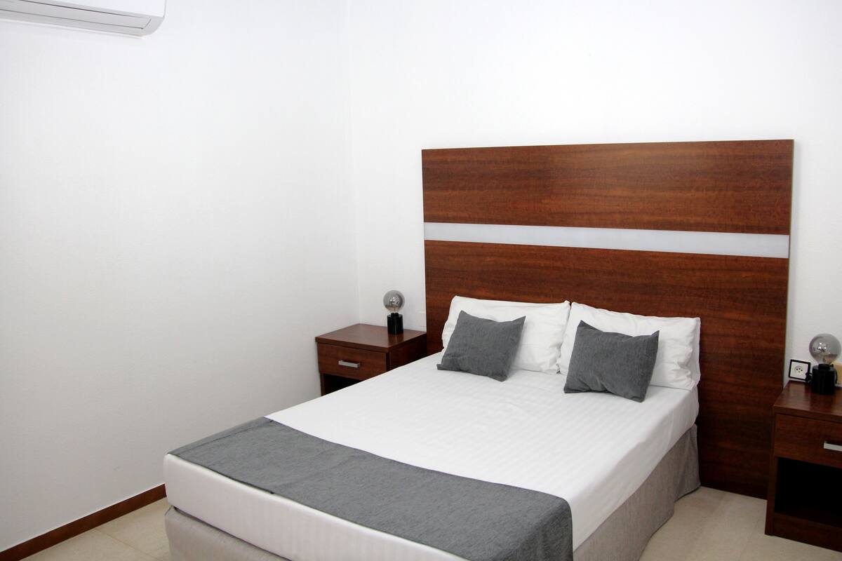 Una cama bien hecha con cabecera de madera, una mesita de noche con lámpara y un aire acondicionado instalado en la pared.