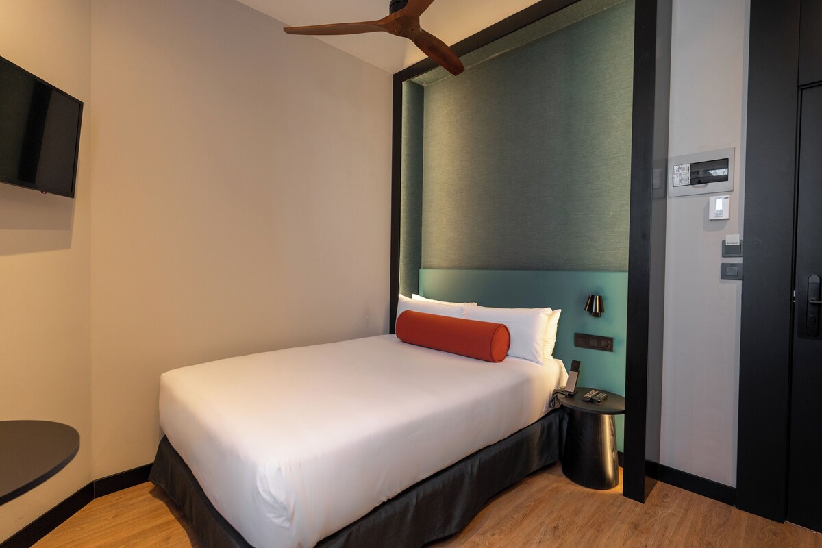 Habitació d'hotel moderna amb un llit individual, una taula de nit, televisió penjada a la paret i un ventilador de sostre.