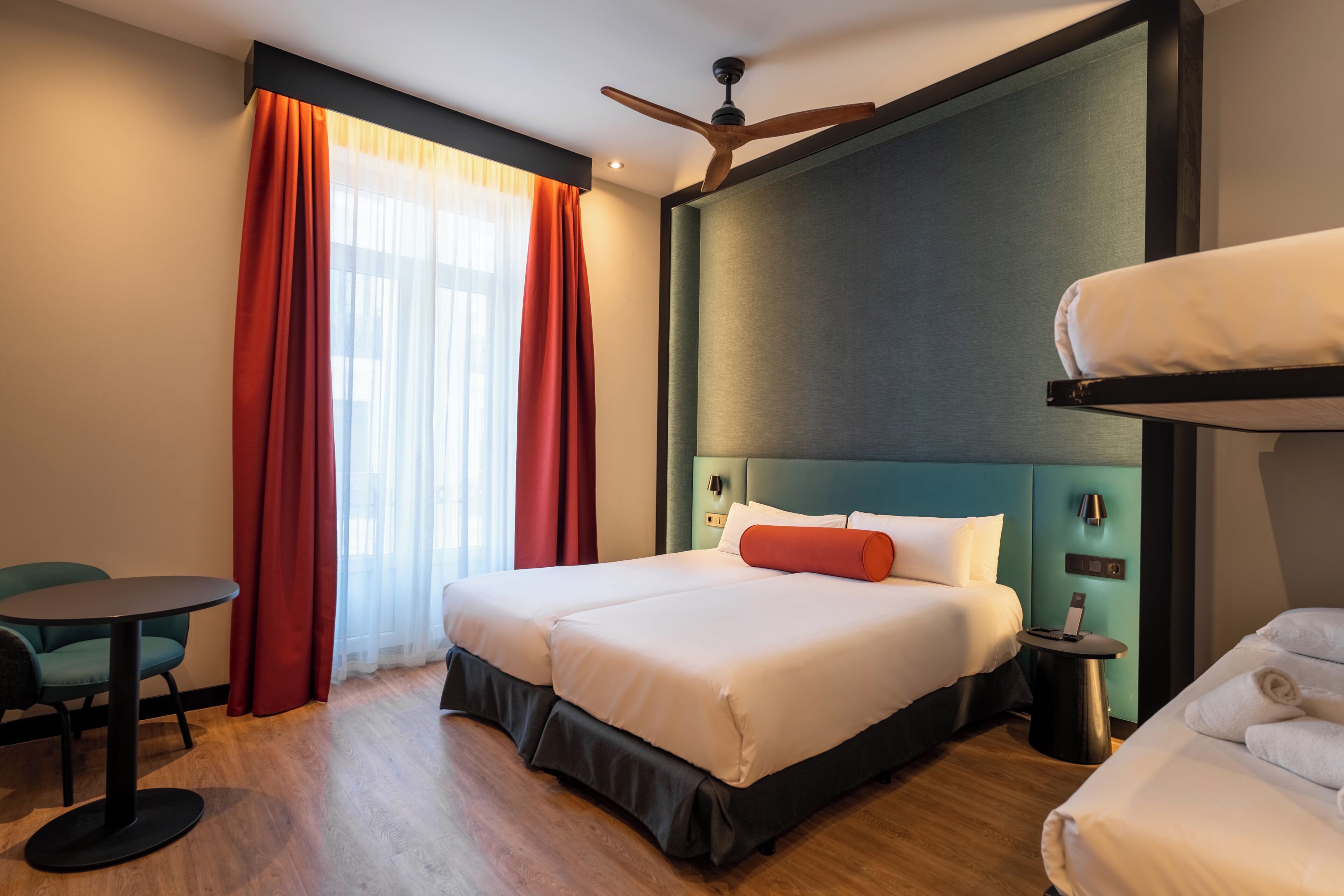 A hotel room with a bed, a desk, a chair, a ceiling fan, and a window with curtains.
