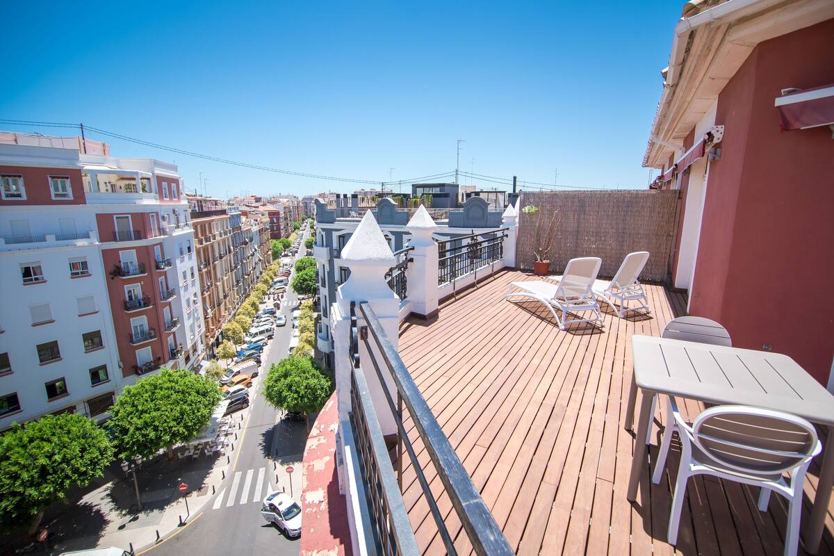 A balcony with white chairs and a table, overlooking a street with parked cars and buildings.