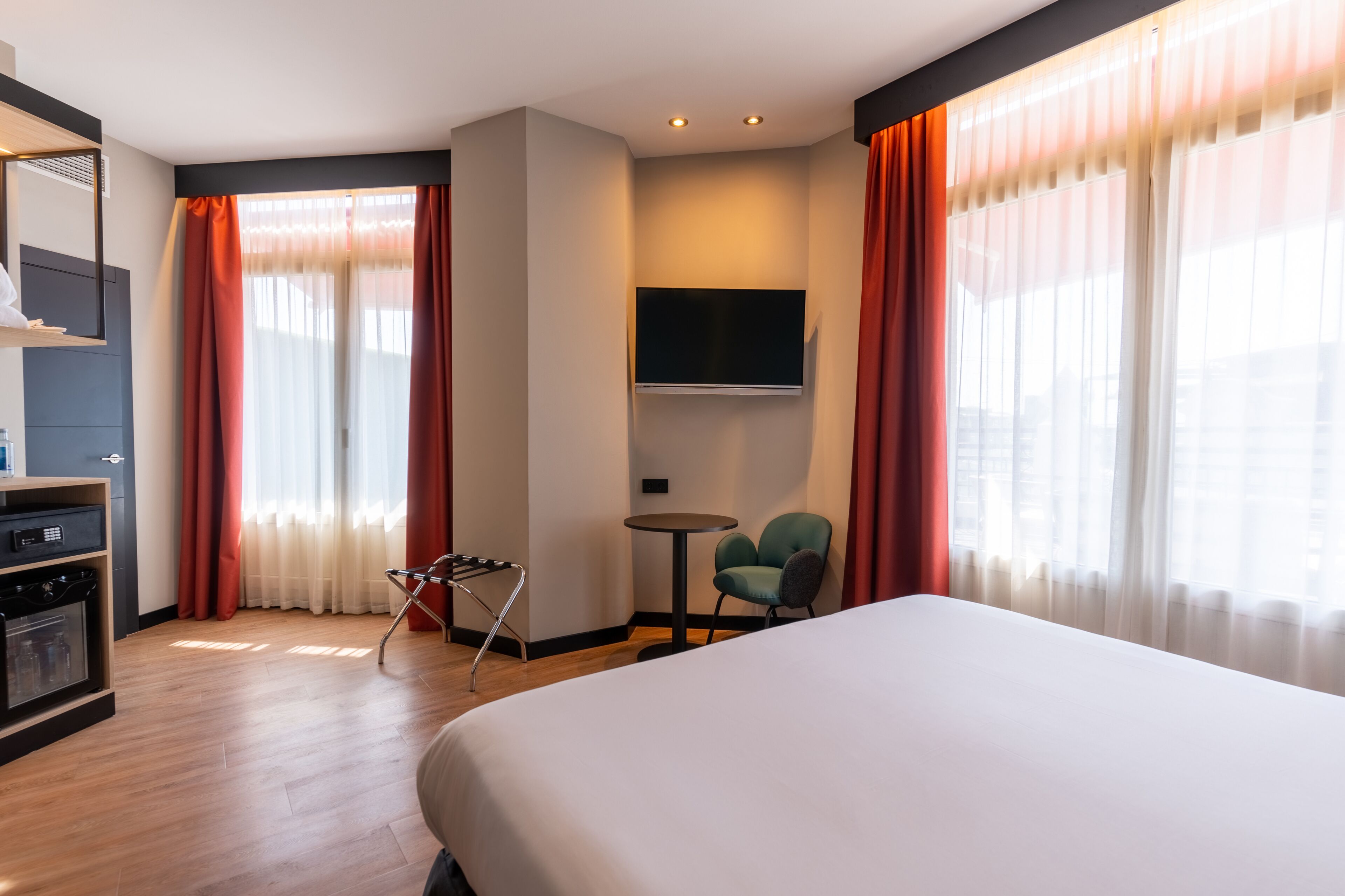 A hotel room with a large bed, a TV mounted on the wall, a small table with a chair, and a minibar.