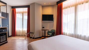 A hotel room with a large bed, a TV mounted on the wall, a small table with a chair, and a minibar.