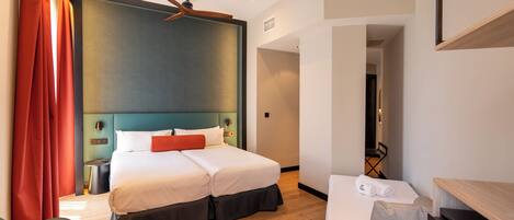 Habitación de hotel con cama, escritorio, silla, ventilador de techo y vista al pasillo.