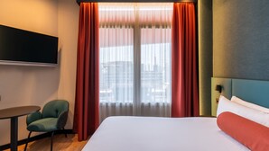 Habitación de hotel con cama, televisor montado en la pared, una silla y ventana con cortinas.