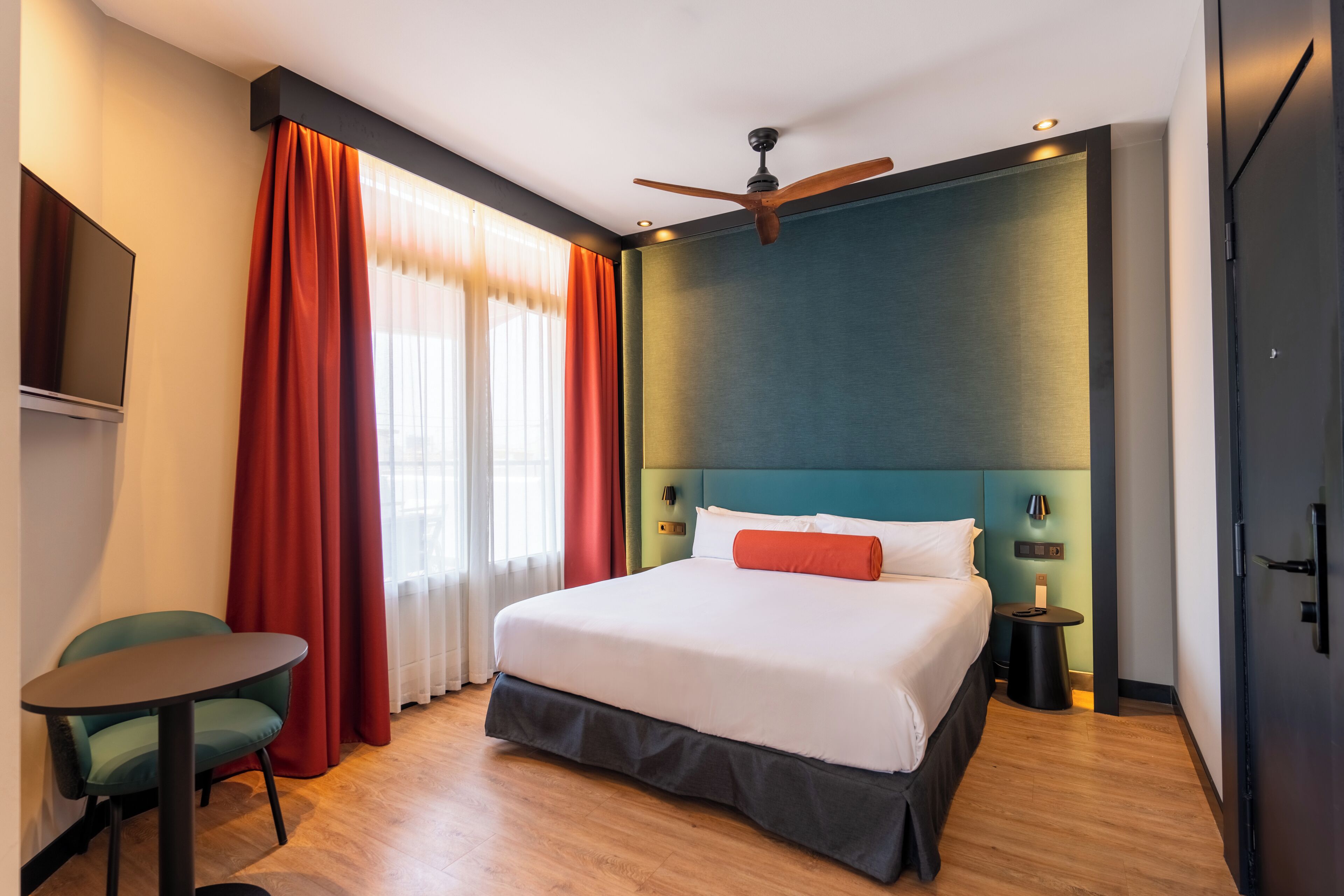 Quarto de hotel com cama, escrivaninha, cadeira, ventoinha no teto e televisão.