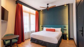Quarto de hotel com cama, escrivaninha, cadeira, ventoinha no teto e televisão.