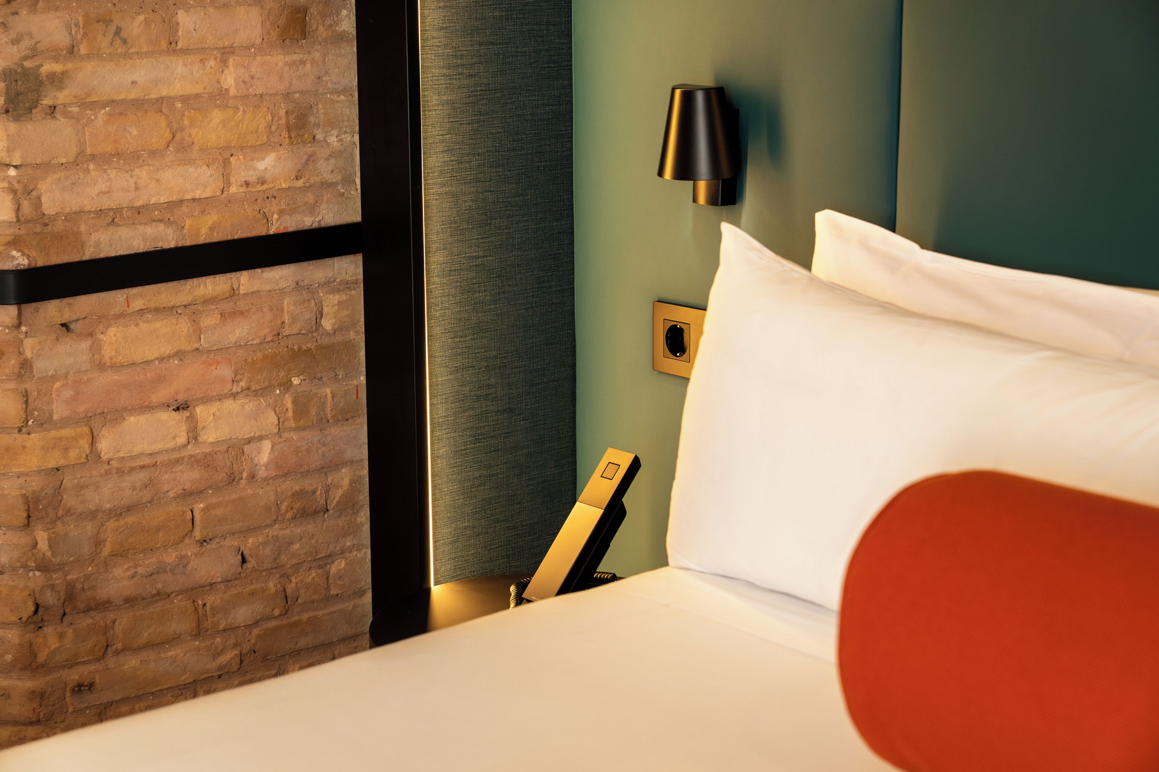 Een hotelkamer met een bakstenen muur, een bed met wit beddengoed, een rode sierkussen, een zwarte lamp en een lichtschakelaar aan de muur.