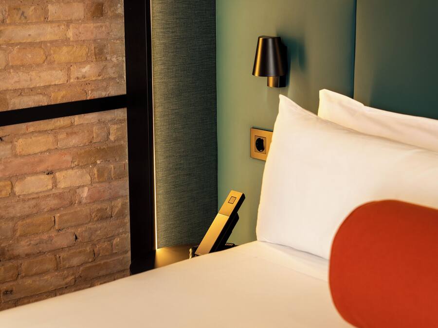 Una habitación de hotel con pared de ladrillo, cama con ropa blanca, almohada decorativa roja, lámpara negra e interruptor de luz en la pared.