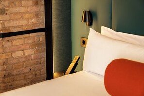 Una habitación de hotel con pared de ladrillo, cama con ropa blanca, almohada decorativa roja, lámpara negra e interruptor de luz en la pared.