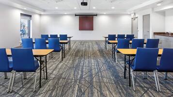Un salón de conferencias con sillas y mesas azules, una pantalla de proyector y piso alfombrado.