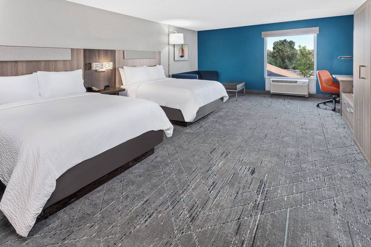 A hotel room with two beds, a desk, a chair, a window with a view, and a wall-mounted air conditioner.
