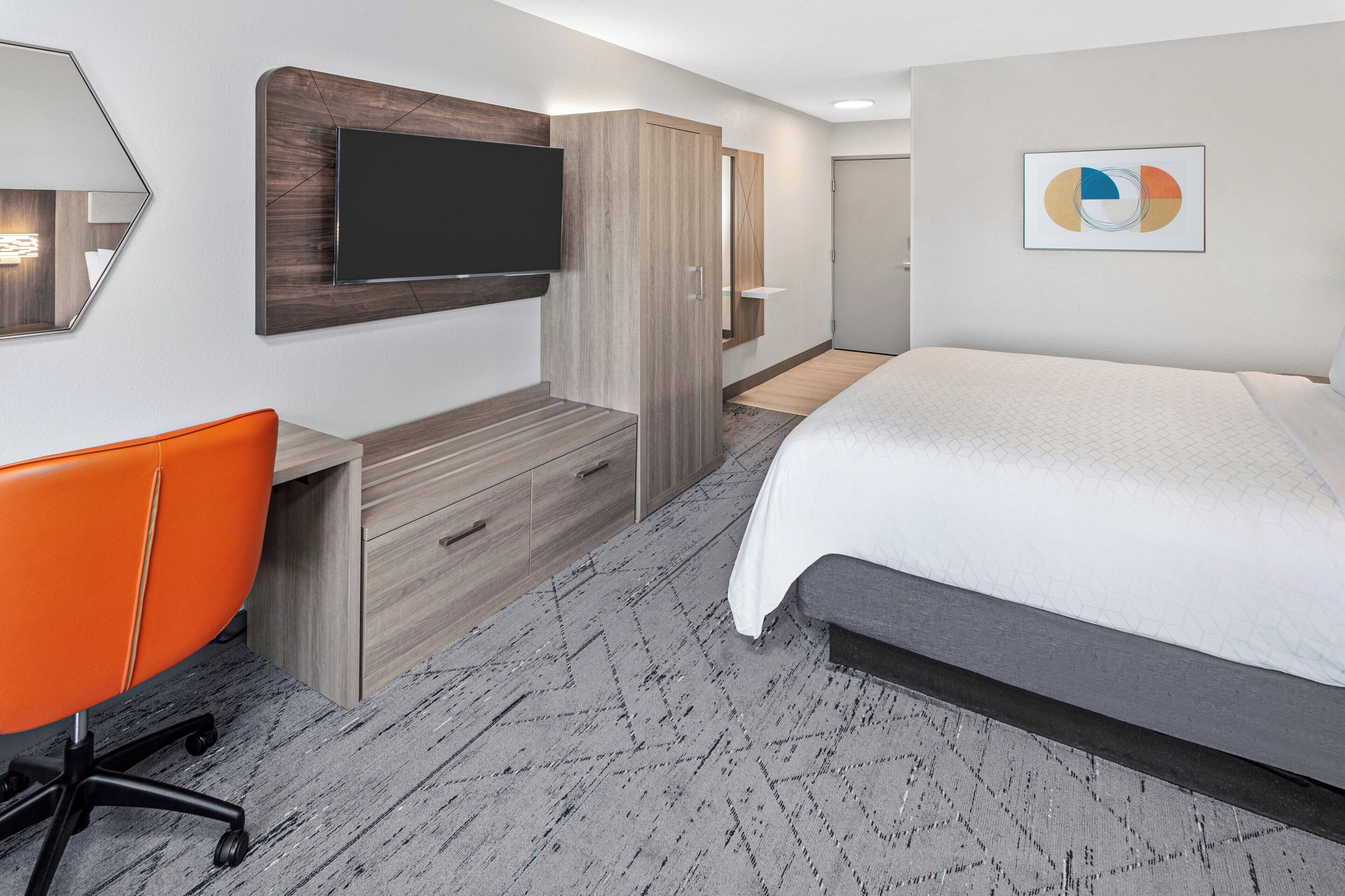 A hotel room with a bed, a desk with a chair, a TV mounted on the wall, and a painting on the wall.