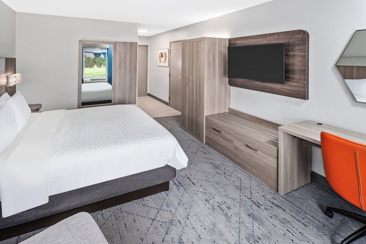 A modern hotel room with a large bed, a desk, a chair, a wardrobe, and a TV mounted on the wall.