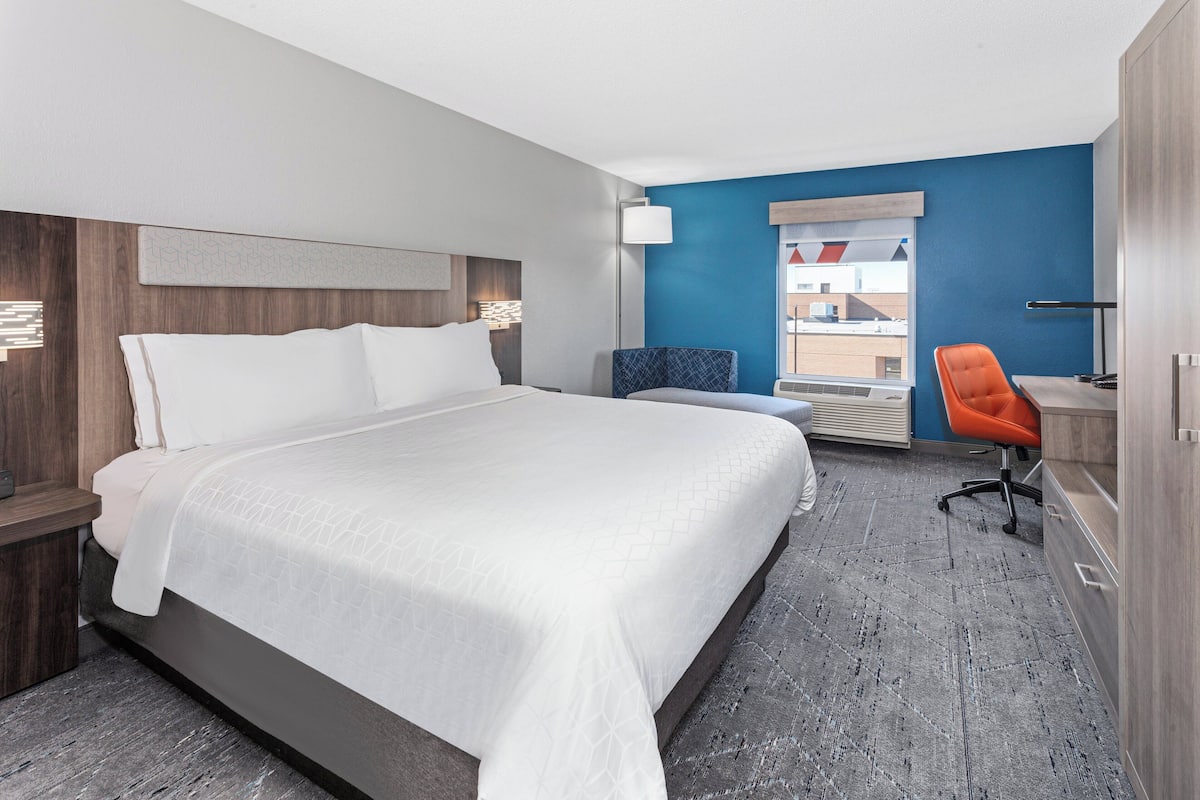 A hotel room with a large bed, a desk with a chair, a window with a view, and a blue accent wall.