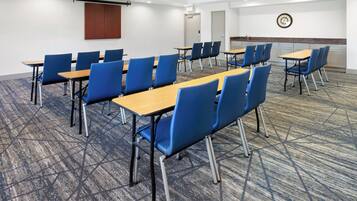A conference room with rows of tables and blue chairs, carpeted floor, white walls, and a clock on the wall.