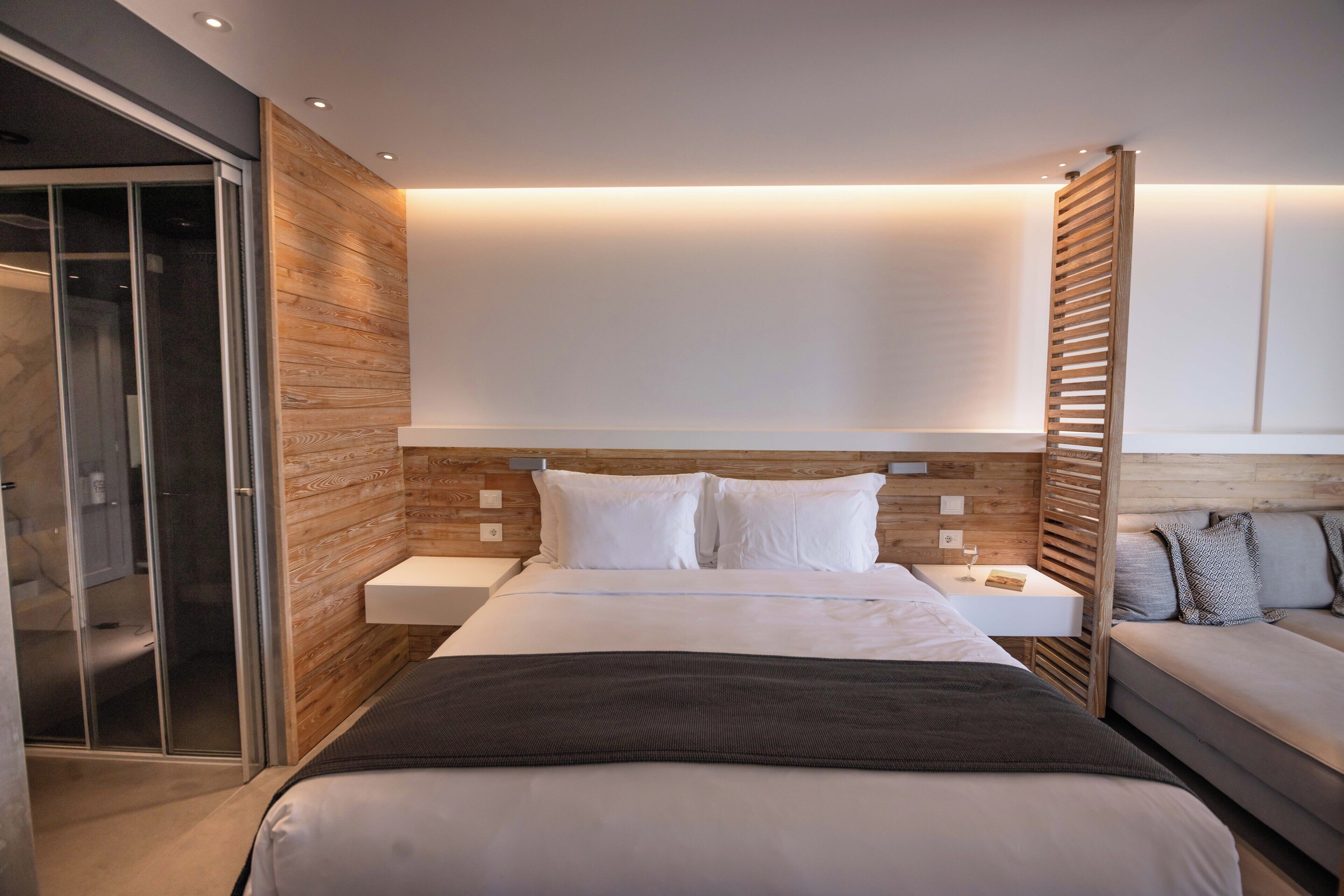 A modern hotel room with a large bed, a bedside table, a built-in wardrobe, and a seating area with a sofa and cushions.