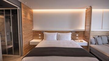 A modern hotel room with a large bed, a bedside table, a built-in wardrobe, and a seating area with a sofa and cushions.