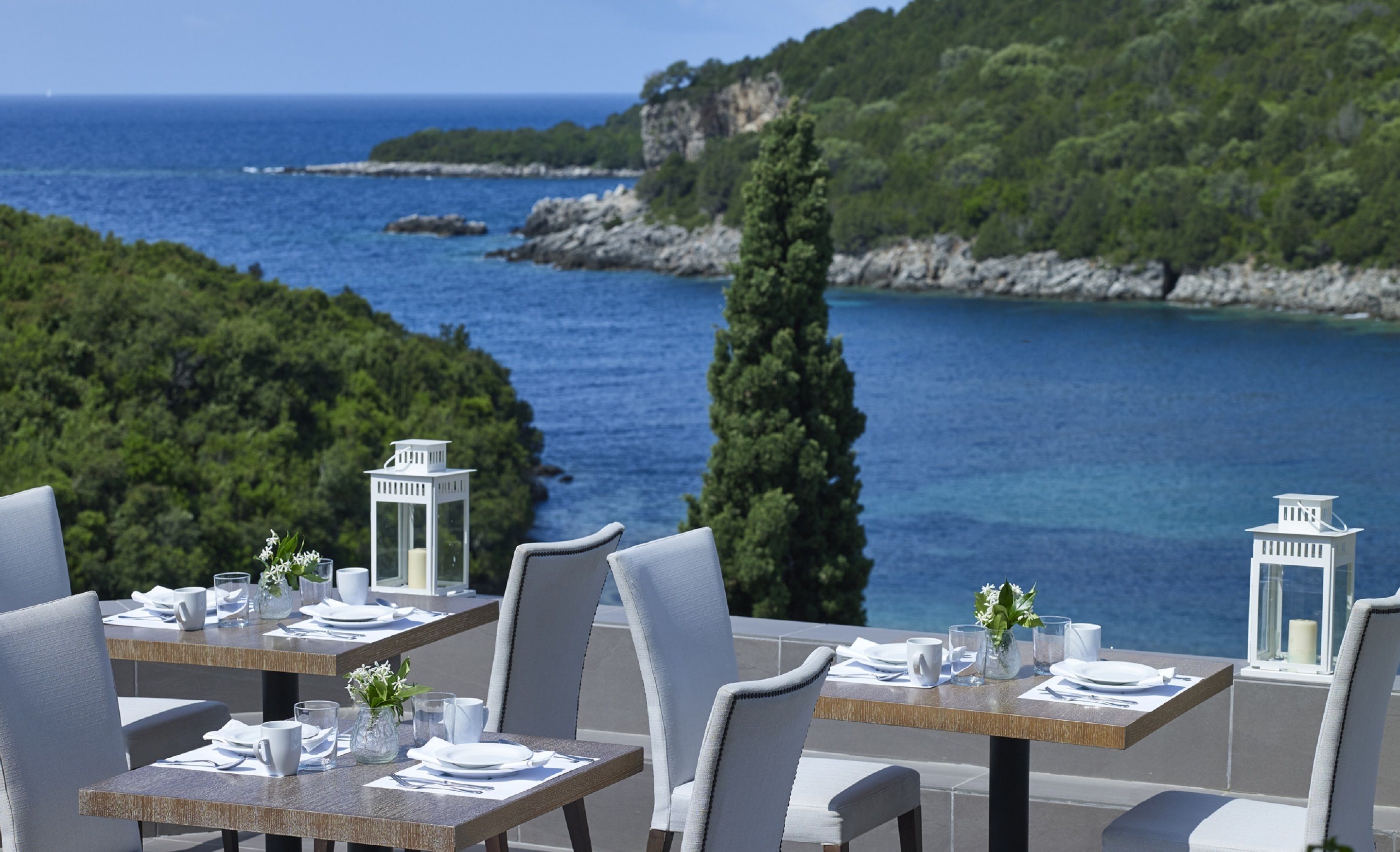 A table set with white tableware and a candle, overlooking a coastal landscape with a clear view of the sea and a rocky island.