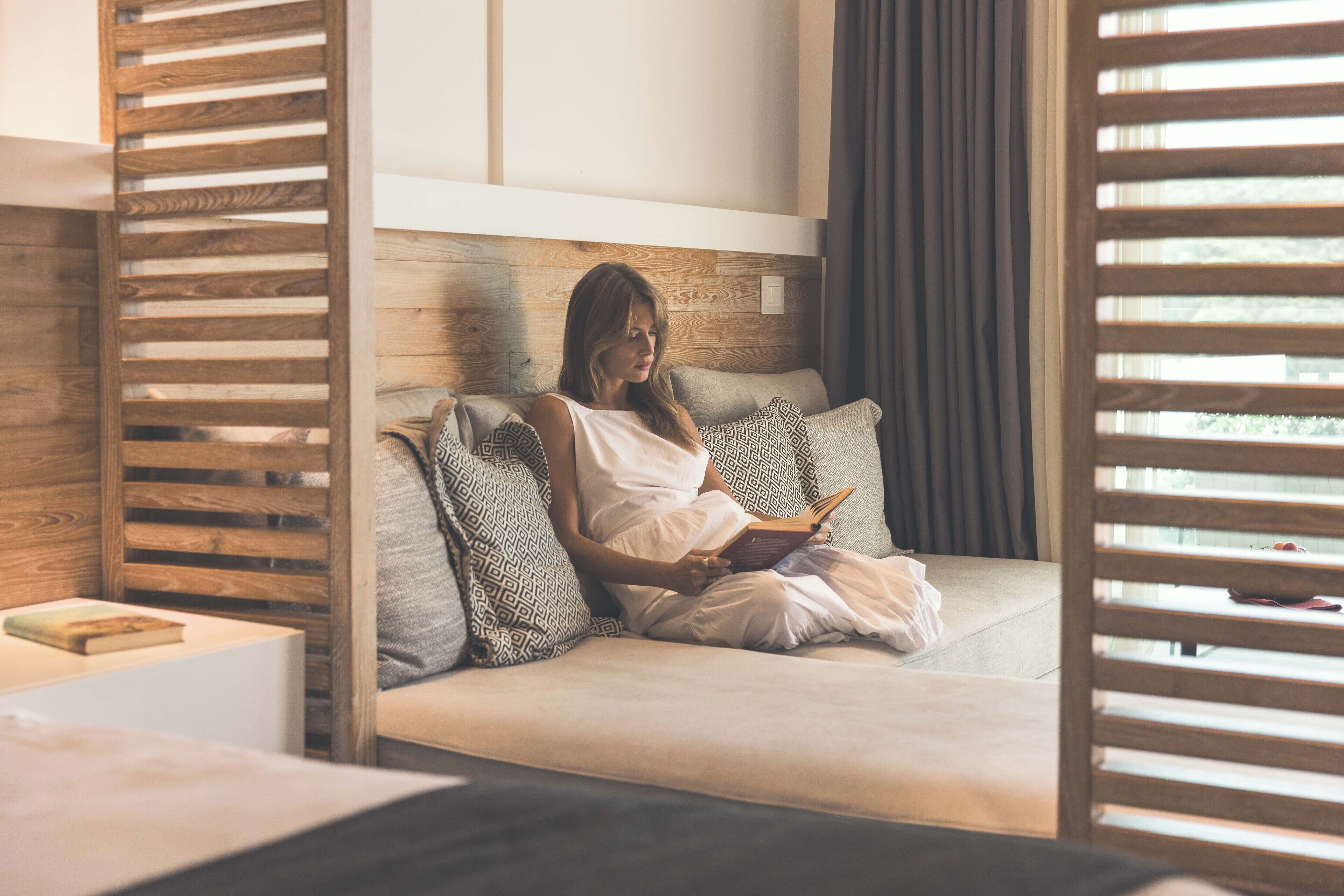 Een vrouw zit op een bank te lezen in een kamer met houten lambrisering en een raam met jaloezieën.