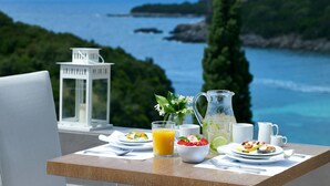 Une table dressée avec des aliments pour le petit-déjeuner, une carafe et un verre de jus d’orange, offrant une vue panoramique sur la côte.