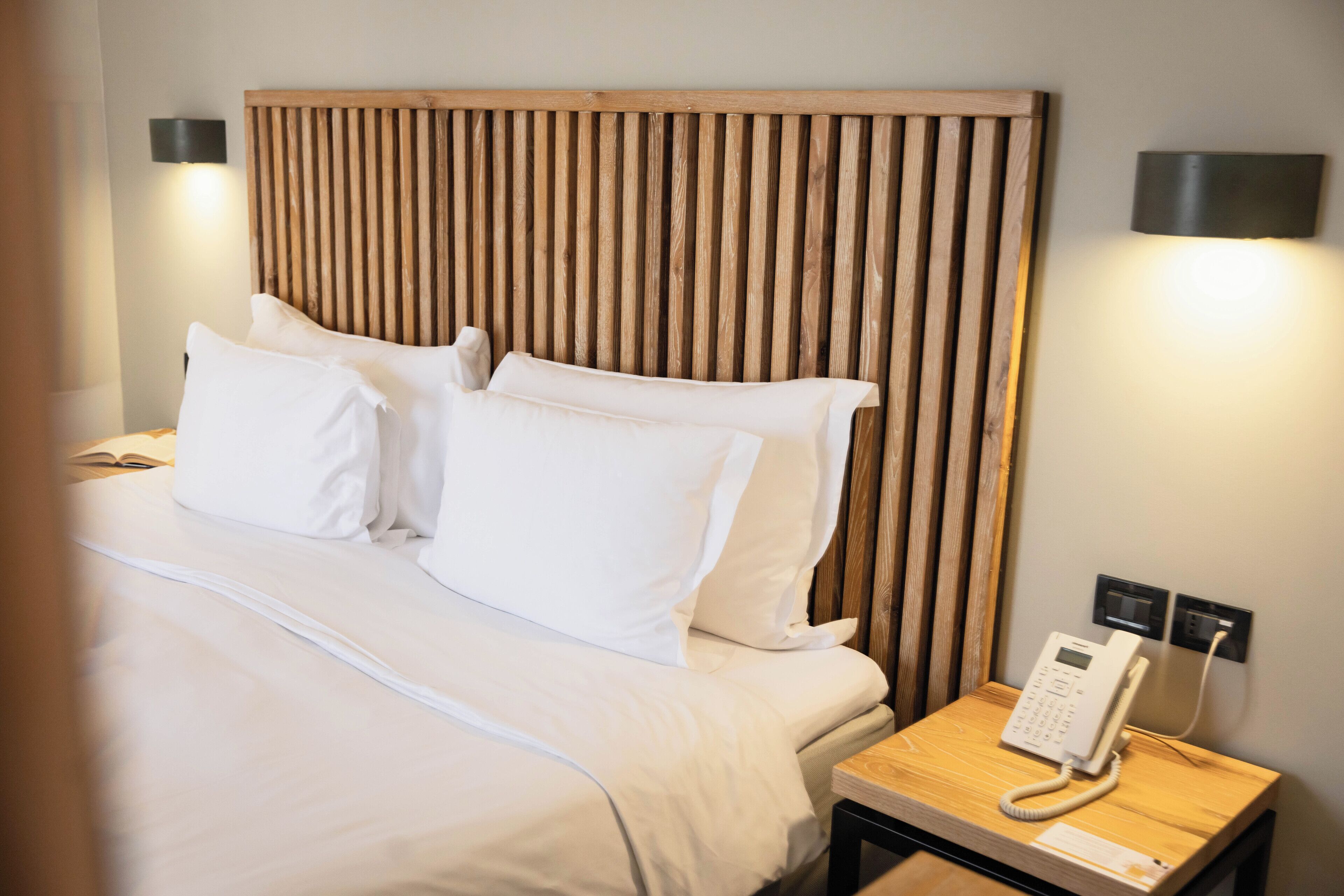 A neatly made bed with white linens and pillows, a wooden headboard, bedside lamps, and a bedside telephone.