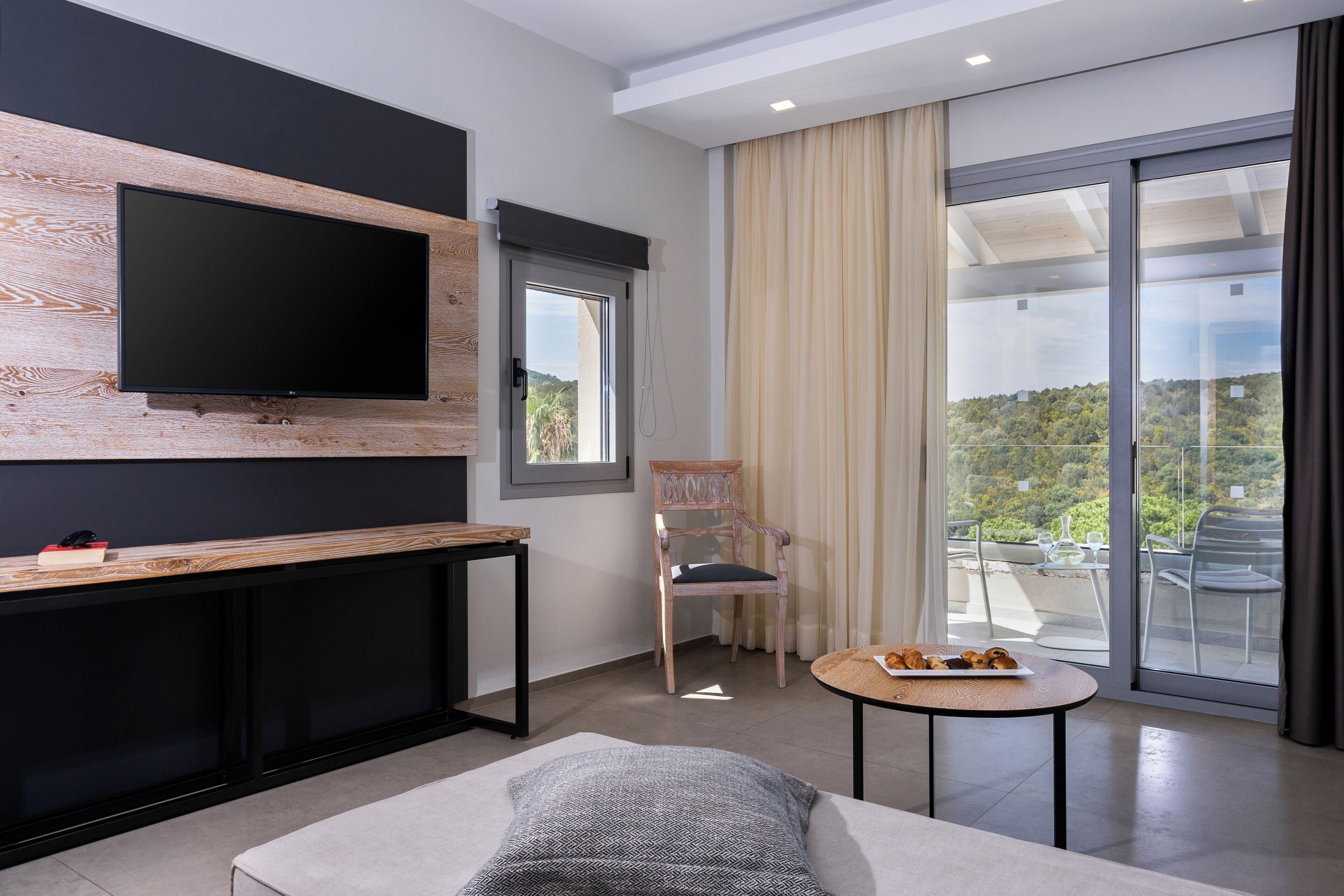 Ein modernes Wohnzimmer mit einem flachbildfernseher an einer Holzwand, einem gemütlichen Sofa, einem kleinen runden Tisch mit Speisen, einem Sessel und einem Balkon mit Blick auf Grünflächen.