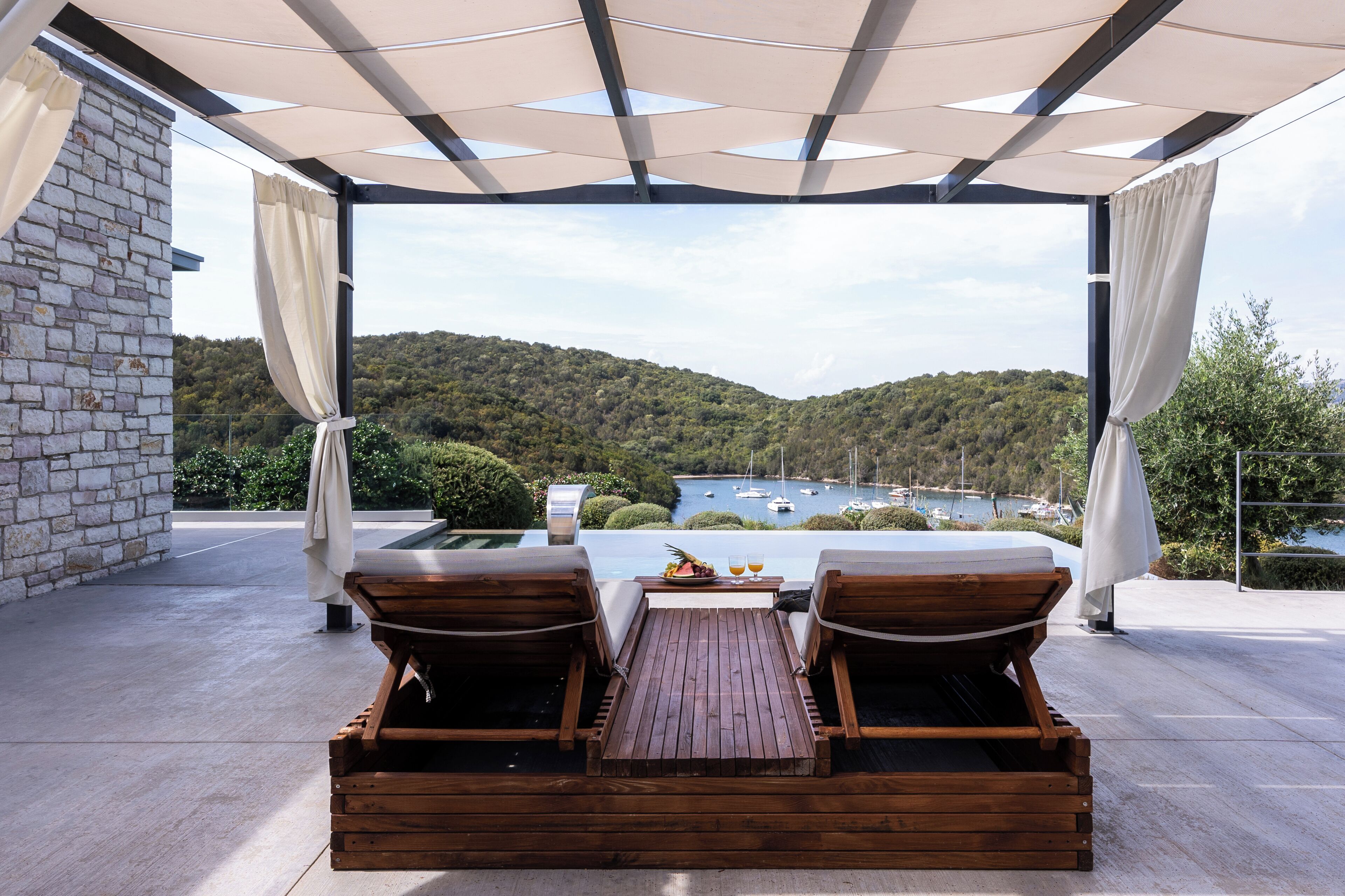 Terraza de madera con dos sillones de descanso y una mesa, con vista a una bahía pintoresca con barcos y colinas.