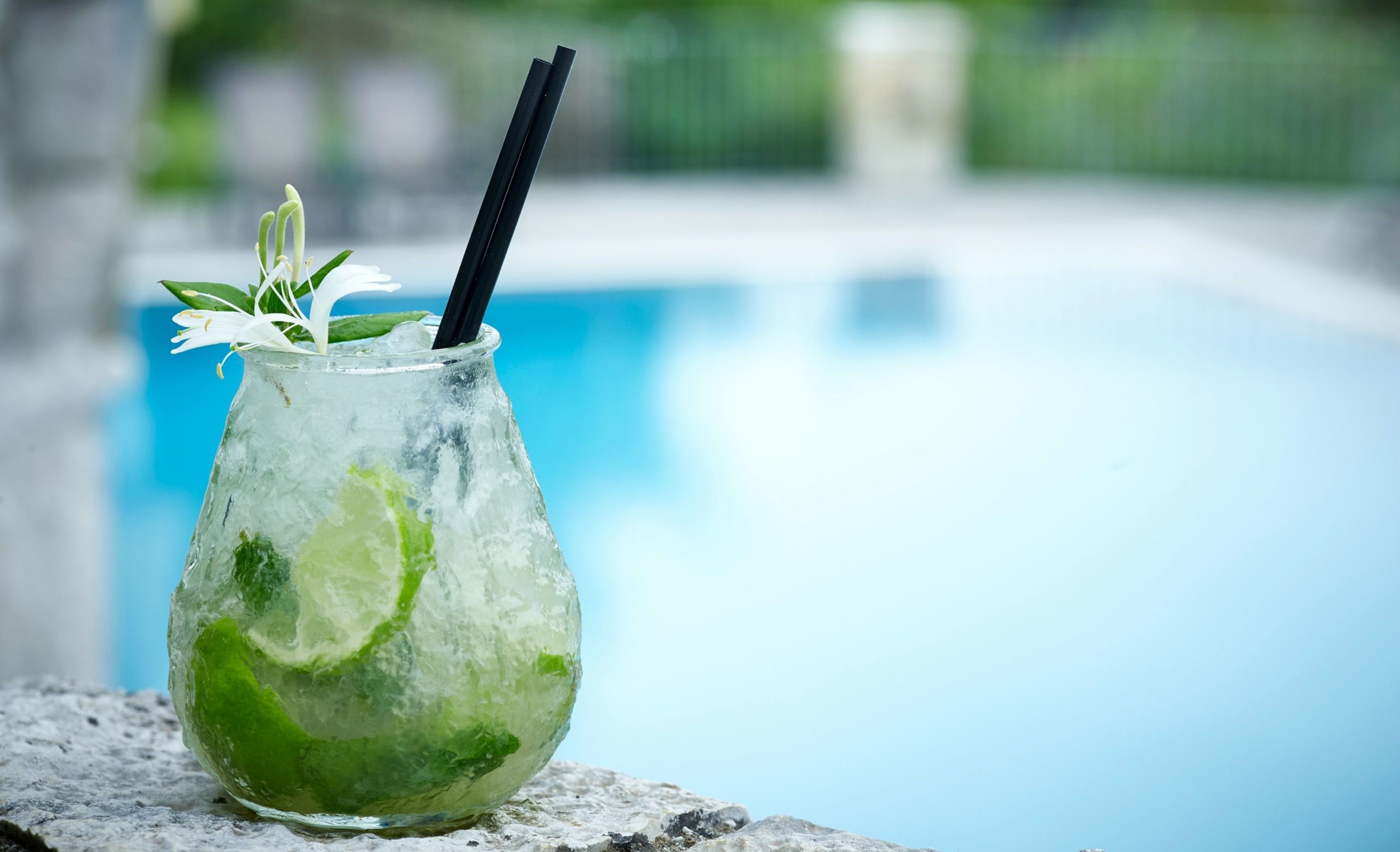 Un vaso de bebida helada con lima y menta, sobre una superficie de piedra cerca de una piscina.
