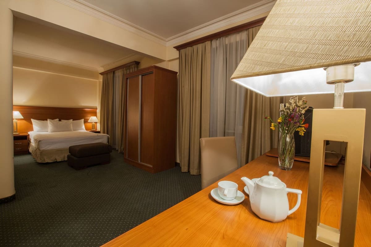Una habitación de hotel con una cama, un escritorio con tetera y taza, una silla, una lámpara y un florero con flores.