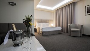 Una habitación de hotel con cama, sofá, sillón, un minibar con champán y dos copas sobre una mesa.