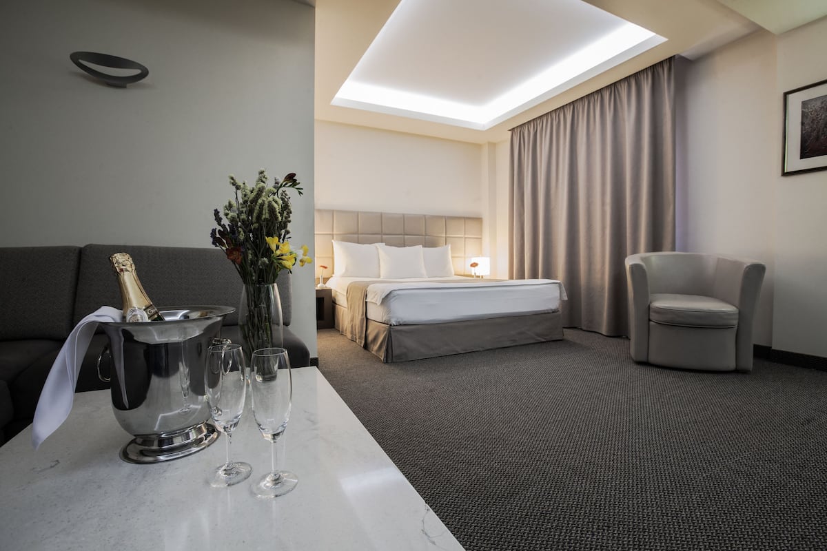 Una habitación de hotel con cama, sofá, sillón, un minibar con champán y dos copas sobre una mesa.
