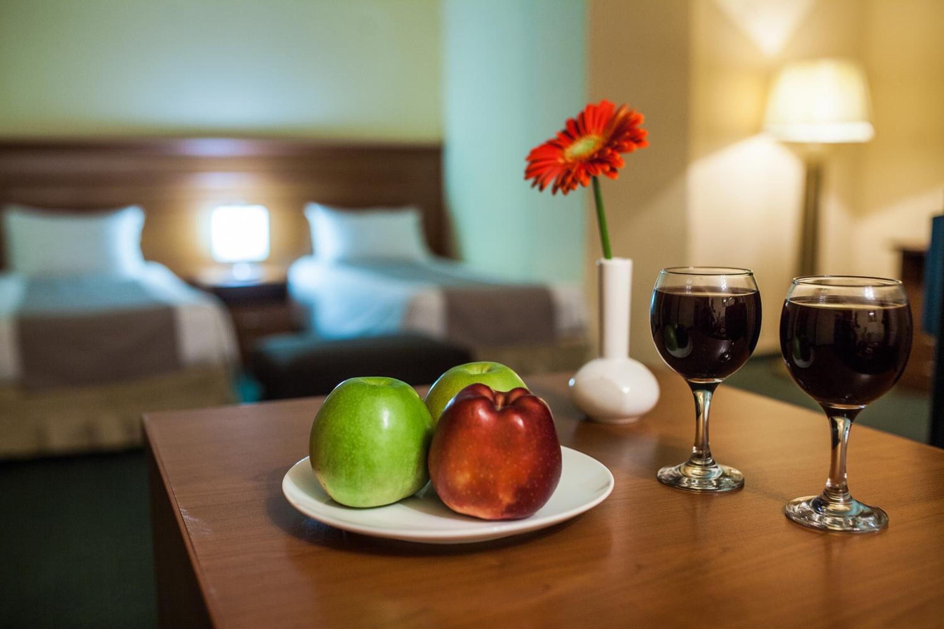 Une chambre d’hôtel avec une tête de lit en bois, un lit recouvert de draps blancs, une petite table sur laquelle se trouve une assiette de pommes, un vase avec une fleur rouge et deux verres à vin.