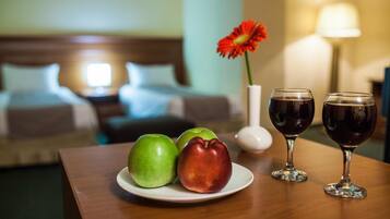Une chambre d’hôtel avec une tête de lit en bois, un lit recouvert de draps blancs, une petite table sur laquelle se trouve une assiette de pommes, un vase avec une fleur rouge et deux verres à vin.
