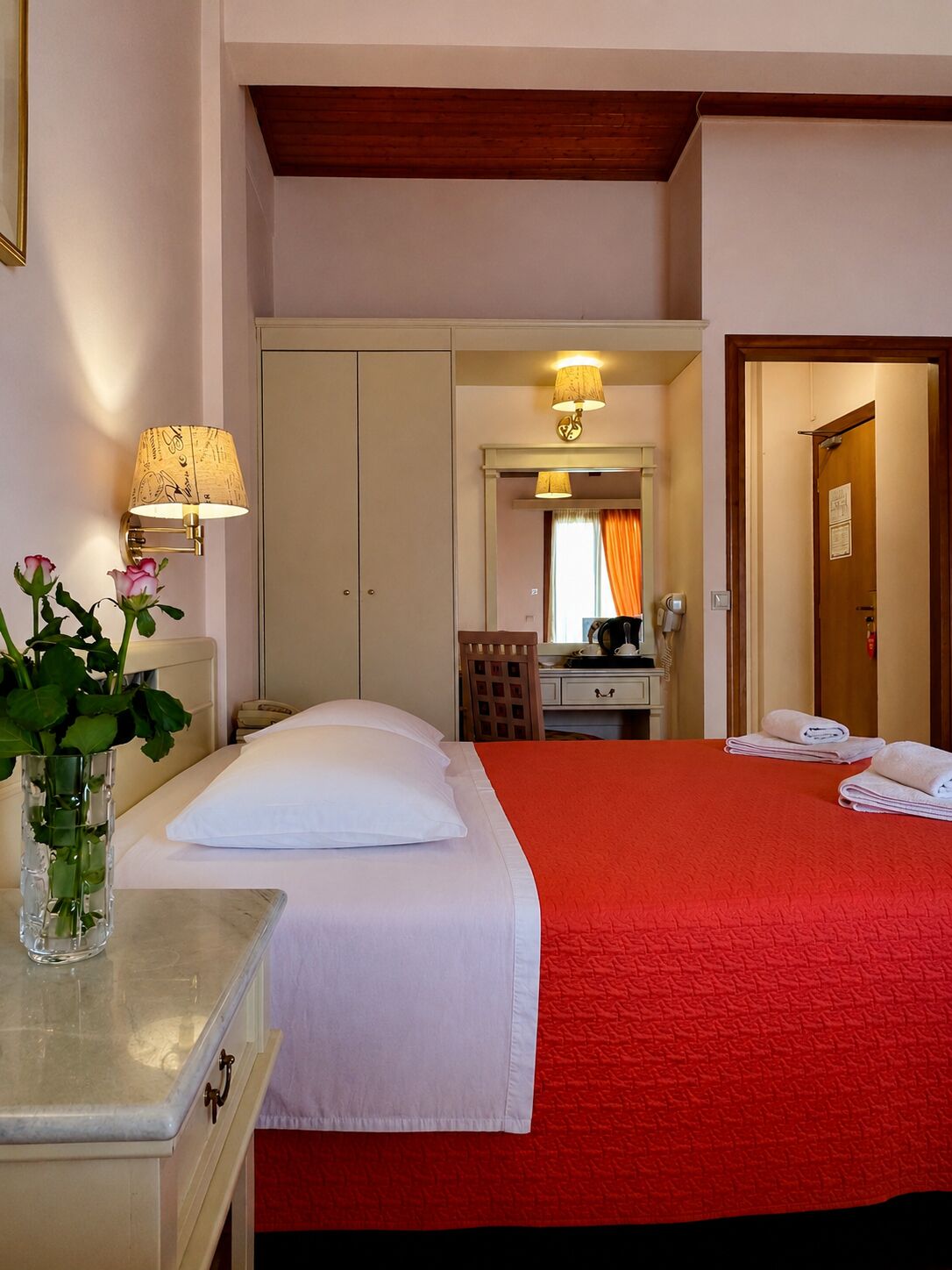 A hotel room with a bed, a desk with a lamp, a mirror, and a door leading to another room.