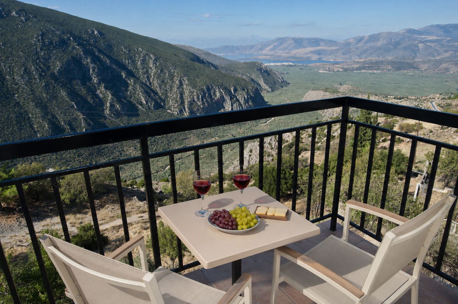 Un balcón con una mesa dispuesta para dos personas, una botella de vino tinto, un bol de uvas y una tabla de quesos.
