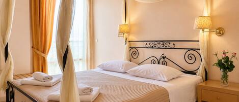 A four-poster bed with a canopy, white linens, two bedside lamps, a nightstand with a vase of flowers, and a window with curtains.
