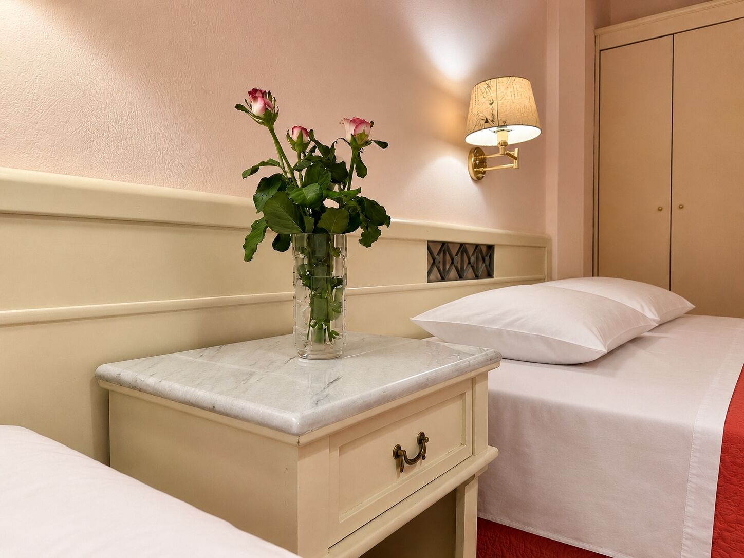 Habitación de hotel con cama, mesita de noche con un jarrón de flores, lámpara de pared y armario.