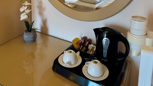 Ein Hotelzimmer mit rundem Spiegel, einem Bett mit weißer Bettwäsche, einer Nachttischlampe, einer kleinen Topf-Orchidee, einer Ablage mit Teetassen und Wasserkocher sowie einer Kaffeemaschine.