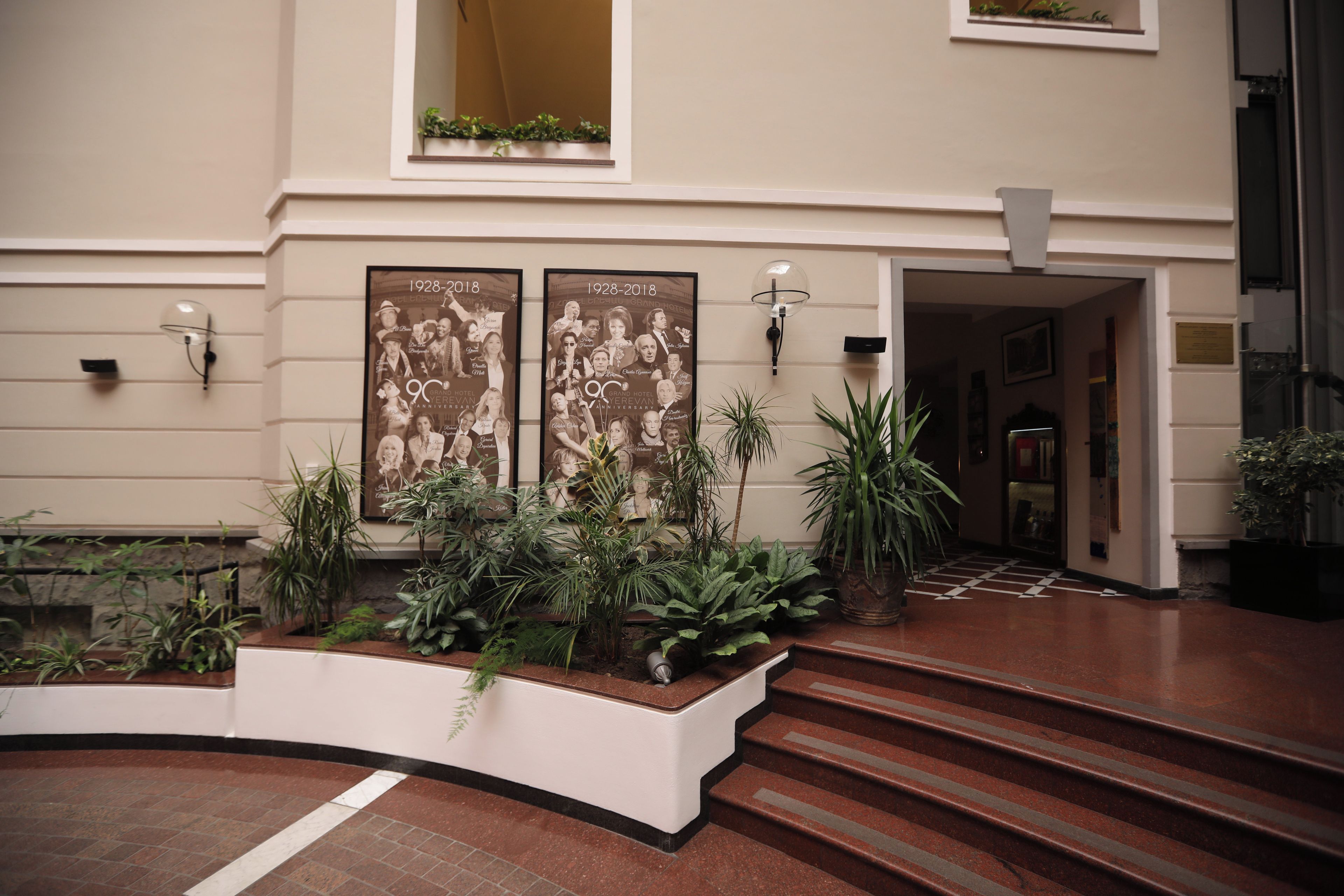 A hotel lobby with a grand staircase, two framed posters, and a variety of indoor plants.